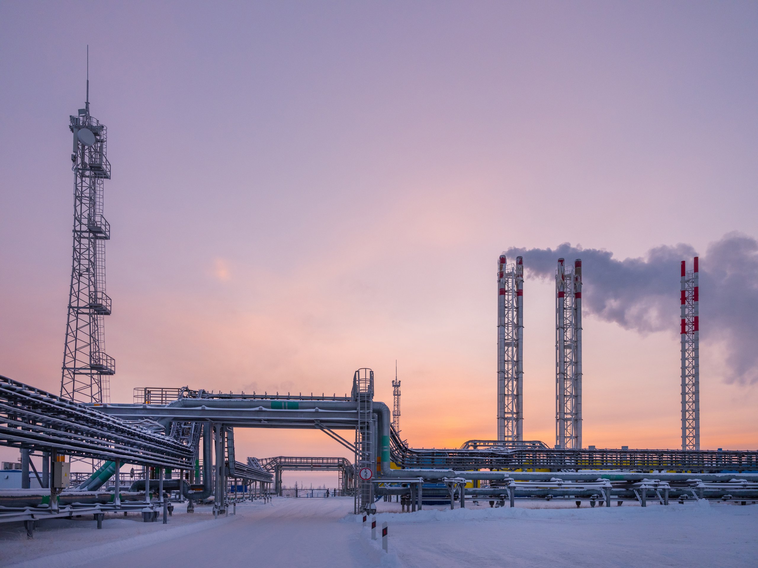 Pipes and steel towers at a natural gas plant are silhouetted against a vibrant, snow-covered sunset landscape during a cold winter evening.